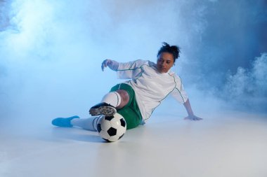 Beyaz ve yeşil elbiseli bayan sporcu dumanın ortasında futbol hareketini sergiliyor.
