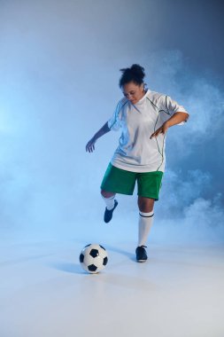 Beyaz formalı ve yeşil şortlu bayan sporcu mavi ışıklı stüdyoda futbol topunu tekmeliyor..