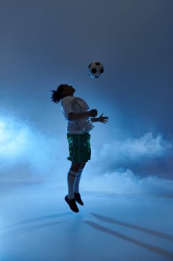Atletik giysili bir kadın mavi arka planda siluetlenir ve bir futbol topunun başına atlar..