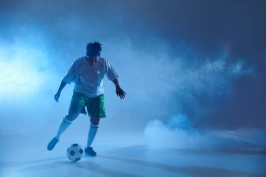 Beyaz formalı bir kadın mavi stüdyoda futbol topu sürüyor..