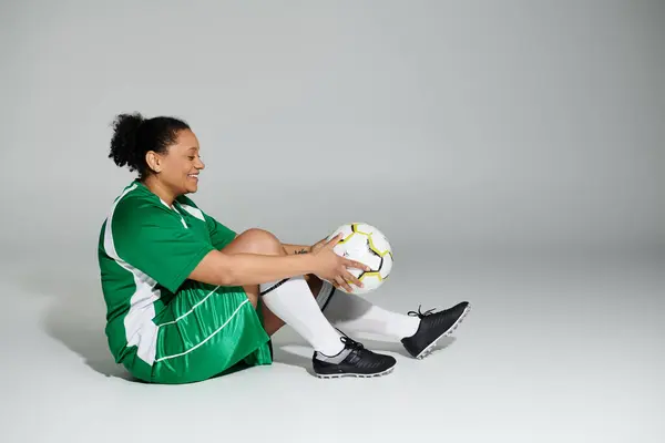 A woman in a green and white soccer uniform sits with a soccer ball.