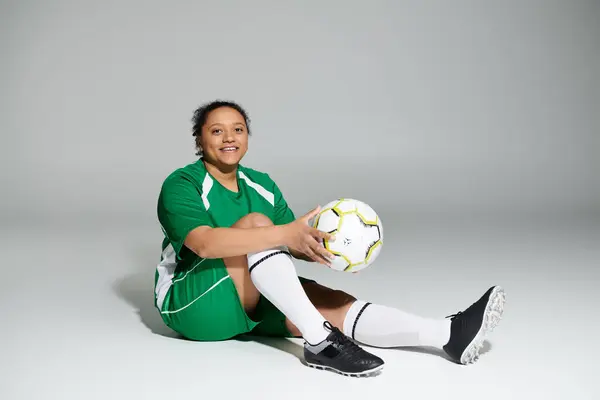 A female soccer player in a green jersey sits on a white studio floor, holding a soccer ball.