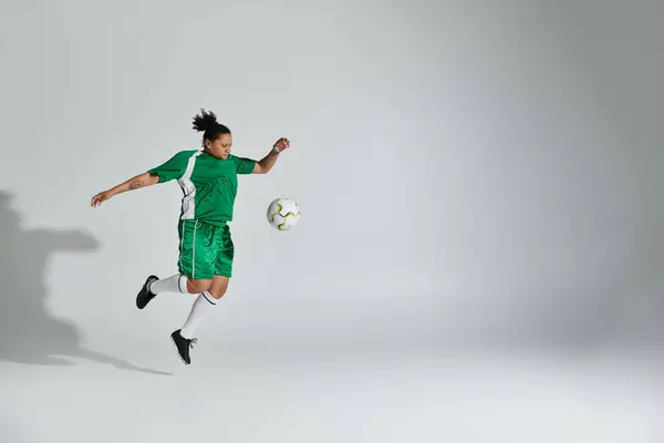 A woman wearing a green jersey and shorts juggles a soccer ball in a studio setting.
