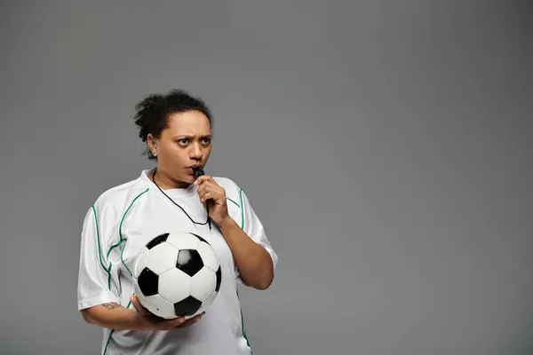 A female referee blows her whistle while holding a soccer ball.