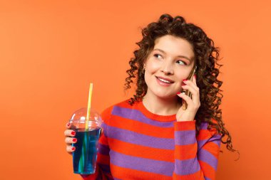 A young woman with curly hair in a vibrant orange and purple striped sweater talks on her phone while holding a blue drink with a straw. clipart