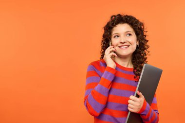 A young woman with curly hair smiles as she talks on her phone and holds a laptop against a vibrant orange background. clipart