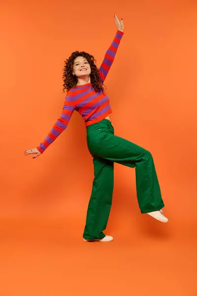 stock image A young woman with curly hair poses in a red and purple striped shirt and green pants against a bright orange background.