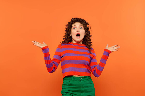 A young woman with curly hair is standing in front of a bright orange background wearing a red and purple striped sweater and green skirt.