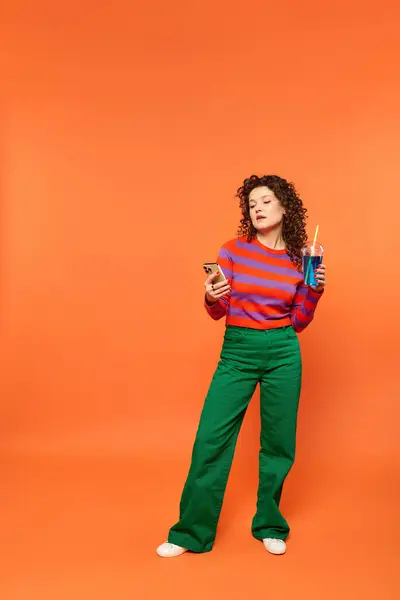 Stock image A young woman with curly hair poses in a colorful outfit against a bright orange background, holding a phone and a blue drink.