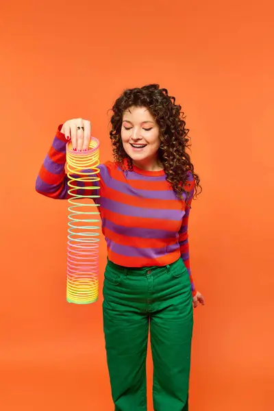 stock image A young woman with curly hair poses against a bright orange background while holding a colorful slinky.