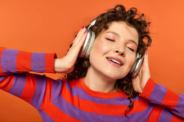 Кудрявая женщина в полосатом свитере, слушает музыку в наушниках на ярко-оранжевом фоне.