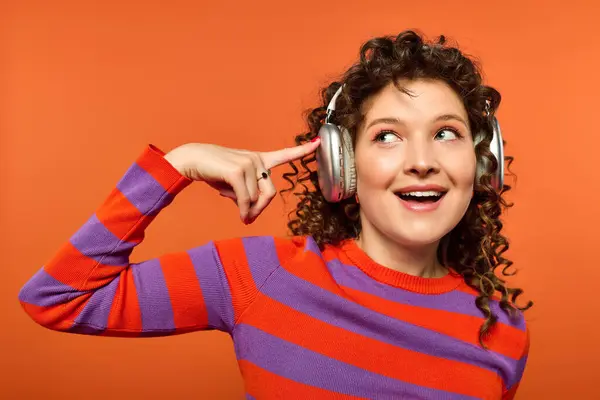 stock image A young woman with curly hair wears vibrant clothing and headphones, smiling brightly against an orange background.