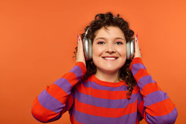 Young woman in striped sweater and headphones smiles against orange background.