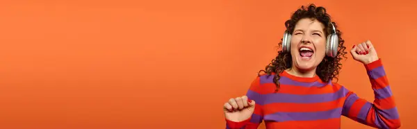 stock image A young woman with curly hair dances enthusiastically while wearing headphones against a bright orange background.