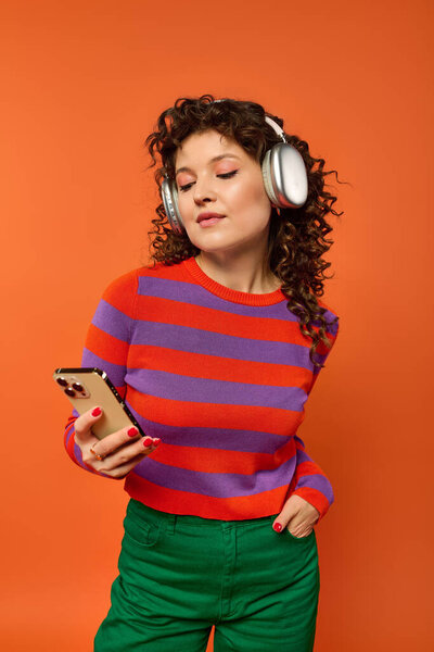 Молодая женщина с вьющимися волосами позирует в ярком одеянии на оранжевом фоне. Она носит наушники и держит смартфон..