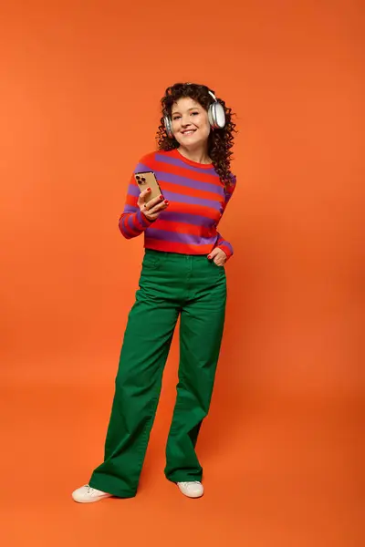 stock image A young woman with curly hair poses in a vibrant red and purple striped sweater and green pants against a bright orange backdrop.
