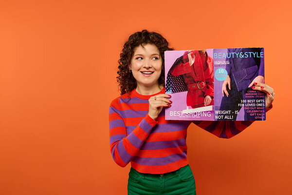 Curly-haired woman holds magazine in front of orange background, wearing striped sweater and green pants.
