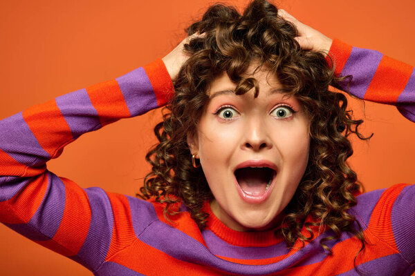 A young woman with curly brown hair poses against a bright orange background, wearing a red and purple striped sweater.