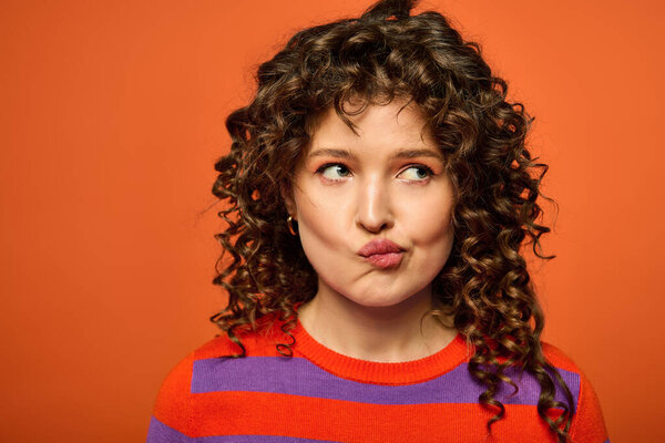 Curly-haired woman in striped sweater poses against orange background, appearing contemplative.