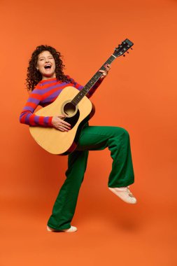 Kıvırcık saçlı ve canlı kıyafetli genç bir kadın parlak turuncu bir arka planda akustik gitar çalıyor..