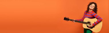 Kıvırcık saçlı genç bir kadın gitarla poz veriyor, parlak turuncu bir arka plana gülümsüyor..