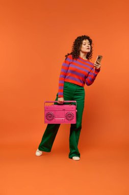 Kıvırcık saçlı genç bir kadın parlak turuncu arka planda pembe bir teyp tutarken canlı bir şekilde poz veriyor..