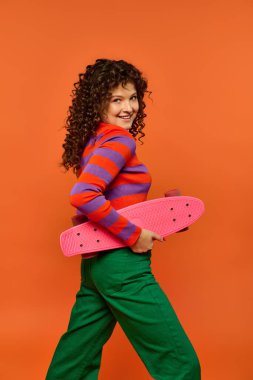 Kıvırcık saçlı, parlak elbiseli, elinde pembe bir kaykayla turuncu arka planda poz veren genç bir kadın..