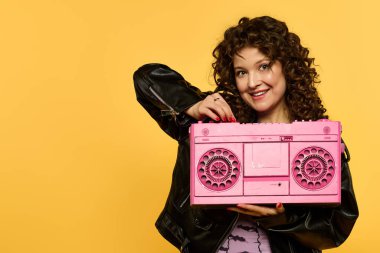 Kıvırcık saçlı, gülümseyen, sarı arka planda pembe teyp taşıyan, siyah deri ceketli bir kadın..