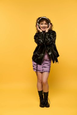 Siyah deri ceketli, sarı arka planda kulaklık takan, kıvırcık saçlı, 20 'li yaşlarda şık bir genç kadın..