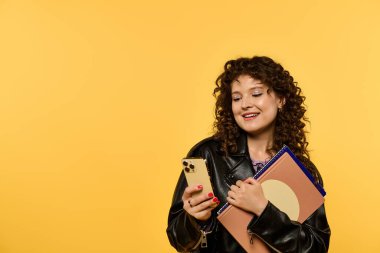 Siyah deri ceket giyen kıvırcık saçlı genç bir kadın elinde defterle telefonuna bakarken gülümsüyor..