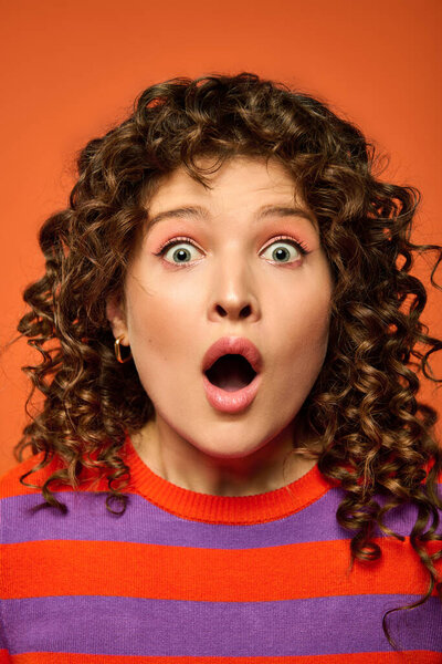 A young woman with curly hair in a striped sweater, mouth open in surprise, against a bright orange background.