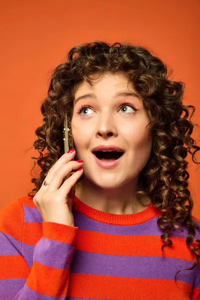 stock image Curly-haired woman in a striped sweater is surprised by a call against bright orange background.