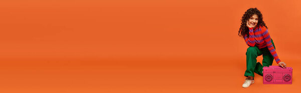 Молодая женщина с вьющимися волосами позирует на оранжевом фоне с розовым бумбоксом. В зеленых штанах и полосатом свитере.