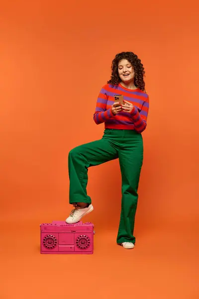 stock image Curly-haired woman in colorful outfit poses against orange background with phone in hand and foot on pink boombox.