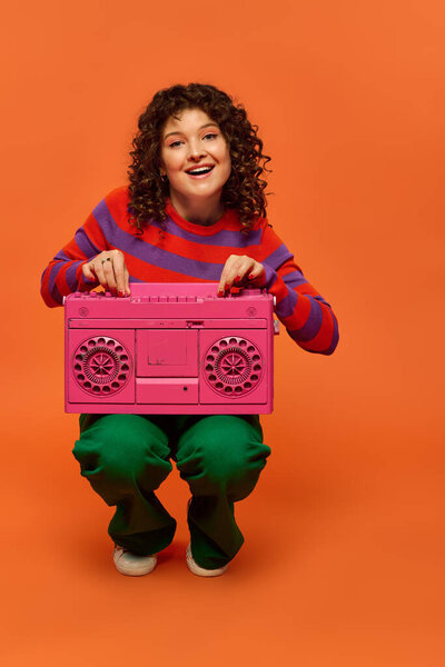 Молодая женщина с вьющимися волосами игриво позирует перед оранжевым фоном, держа розовый бумбокс.