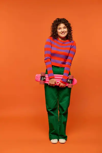 stock image A young woman with curly hair poses with a pink skateboard against a bright orange background.