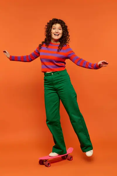 stock image A young woman with curly hair, wearing a striped shirt and green pants, poses on a pink skateboard against a bright orange background.