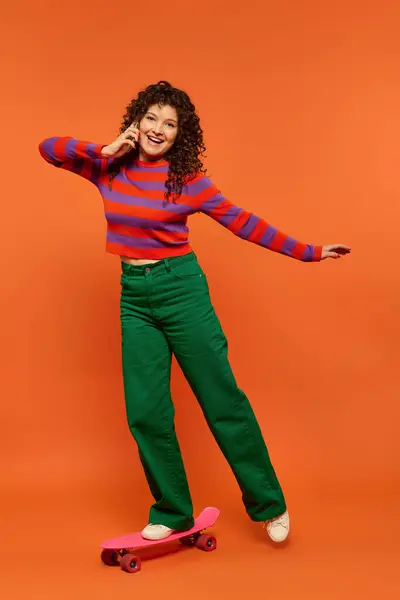 Stock image A young woman with curly hair poses on a pink skateboard against a bright orange background, wearing a striped sweater and green pants.