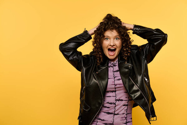 A young woman with curly hair and a black leather jacket poses against a yellow background, looking surprised and excited.