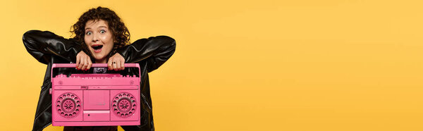 Стильная молодая женщина с вьющимися волосами позирует в черной кожаной куртке, держа розовый бумбокс. Фон яркий желтый.