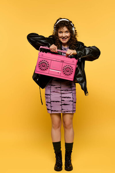 Молодая женщина с вьющимися волосами позирует в черной кожаной куртке, держа розовый бумбокс на жёлтом фоне.