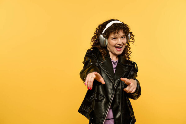 Молодая женщина с вьющимися волосами, в черной кожаной куртке, уверенно позирует с яркой улыбкой, на ярком желтом фоне.