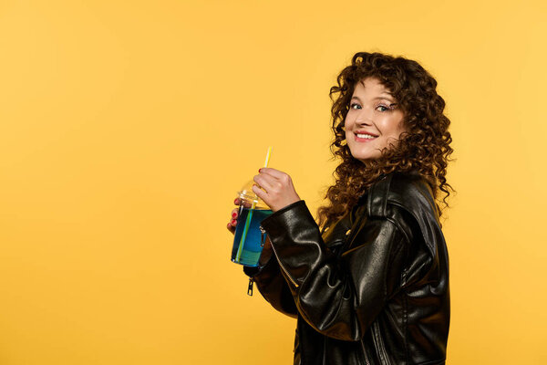 Young woman with curly hair smiles holding a blue drink, styled in a black leather jacket against a bright yellow background.