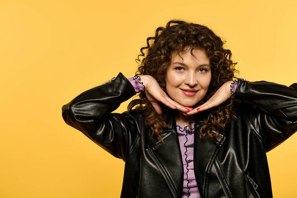 Curly-haired woman in leather jacket smiles and playfully gestures against yellow background.