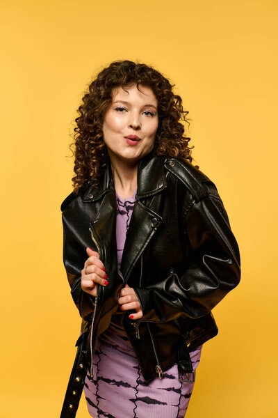 Молодая женщина с вьющимися волосами позирует в черной кожаной куртке на ярком желтом фоне.