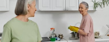 A mature lesbian couple cleans their modern apartment together. One woman washes dishes while the other looks on. clipart