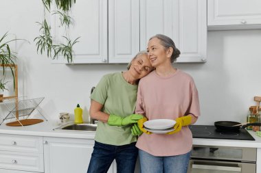 Two mature women, a lesbian couple, stand in their modern kitchen with a happy expression as they complete their household chores. clipart