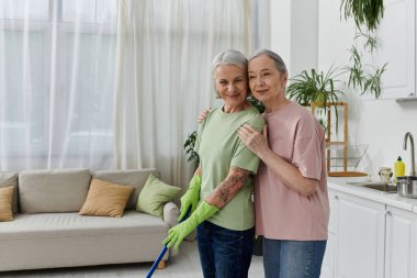 Olgun bir lezbiyen çift, elinde paspas ve yeşil temizleme eldiveni olan bir kadınla birlikte modern dairelerini temizliyor..