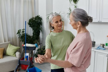 Yaşlı bir lezbiyen çift modern dairelerini birlikte temizliyor, gülüyor ve birbirlerine eşlik ediyorlar..