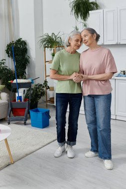 Two mature lesbian women, holding hands, take a break from cleaning their modern apartment. clipart